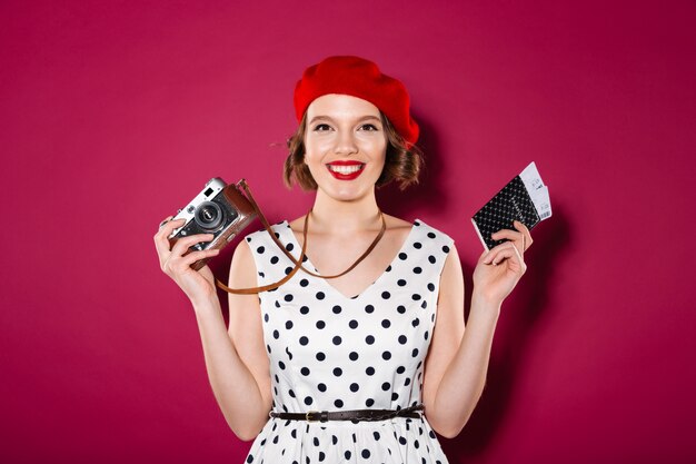 Feliz mujer de jengibre en vestido con pasaporte con entradas y cámara retro mientras mira a la cámara en rosa