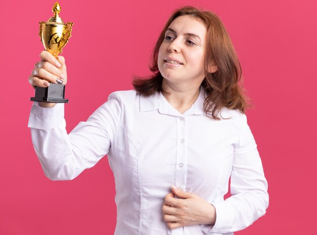 Feliz mujer joven en camisa blanca con trofeo de oro mirándolo con expresión de confianza de pie sobre la pared rosa