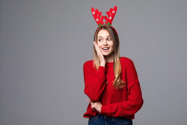 Feliz mujer joven con cuernos de reno en la cabeza mira y apunta a la cámara en la pared gris