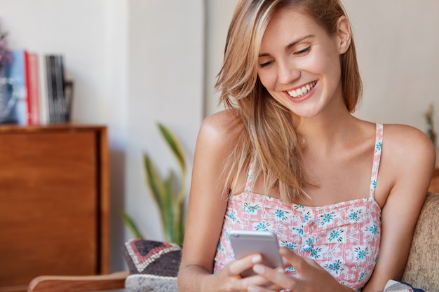 Feliz mujer joven con expresión alegre se sienta en un cómodo sofá, escribe mensajes en el teléfono celular,