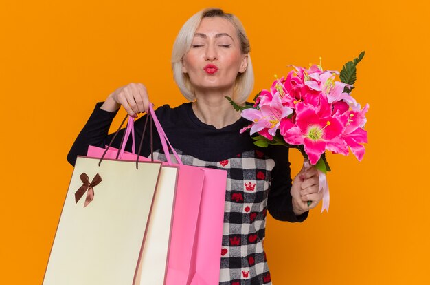 Feliz mujer joven en un hermoso vestido con ramo de flores y bolsas de papel con regalos que soplan un beso celebrando el día internacional de la mujer de pie sobre la pared naranja