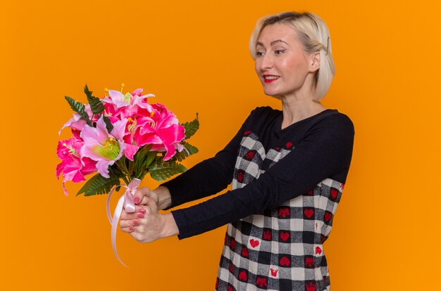 Feliz mujer joven en un hermoso vestido sosteniendo un ramo de flores sonriendo alegremente celebrando el día internacional de la mujer de pie sobre la pared naranja