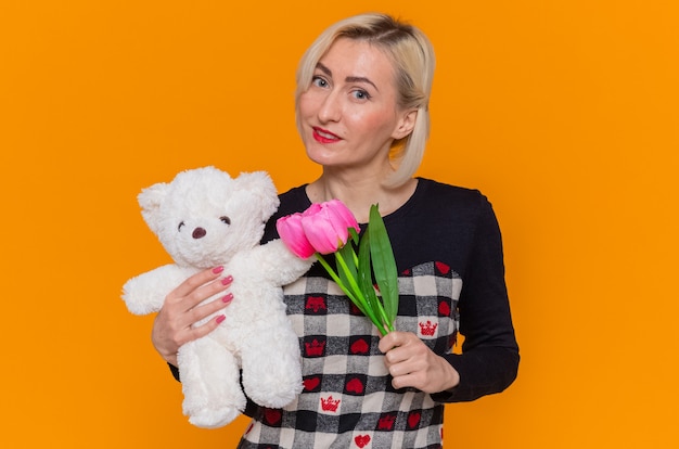 Feliz mujer joven en un hermoso vestido sosteniendo un ramo de tulipanes y un oso de peluche como regalos mirando al frente sonriendo celebrando el día internacional de la mujer de pie sobre la pared naranja