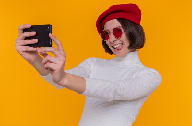 Feliz mujer joven con pelo corto en cuello alto blanco con boina y gafas de sol rojas haciendo selfie con smartphone sonriendo alegremente de pie sobre la pared naranja