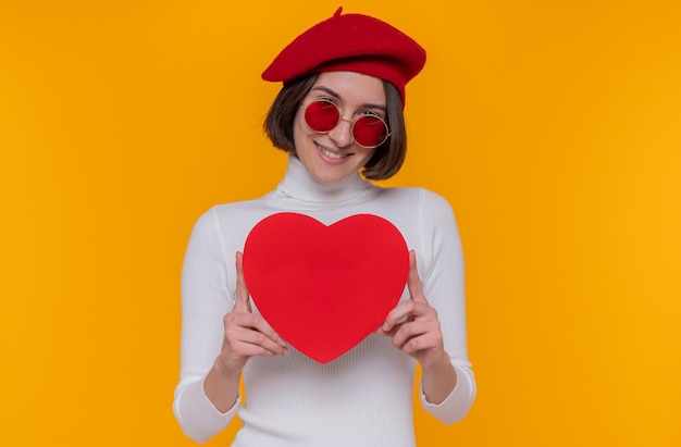 Feliz mujer joven con pelo corto en cuello alto blanco con boina y gafas de sol rojas sosteniendo corazón hecho de cartón mirando al frente sonriendo alegremente parado sobre pared naranja