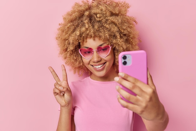 Feliz mujer joven de pelo rizado hace un gesto de paz, se toma una selfie a través de un teléfono inteligente, usa gafas de sol en forma de corazón y una camiseta casual se siente positiva aislada sobre un fondo rosa Toma monocromática