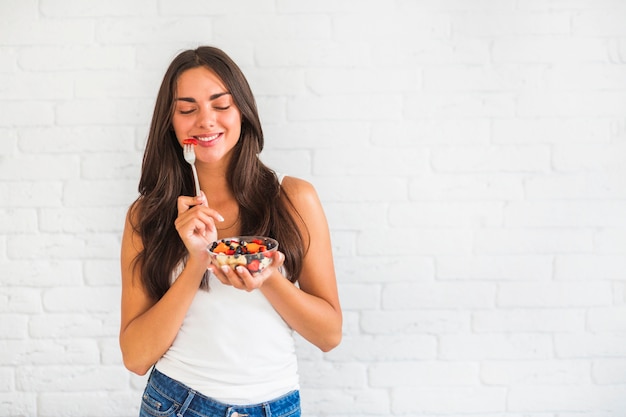 Feliz mujer joven de pie contra la pared blanca comiendo ensalada de frutas