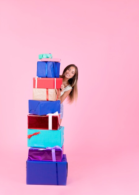 Feliz mujer joven de pie detrás de la pila de cajas de regalo diferentes sobre fondo rosa