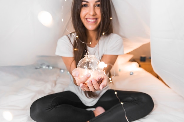 Feliz mujer joven sentada en la cama sosteniendo esfera brillante