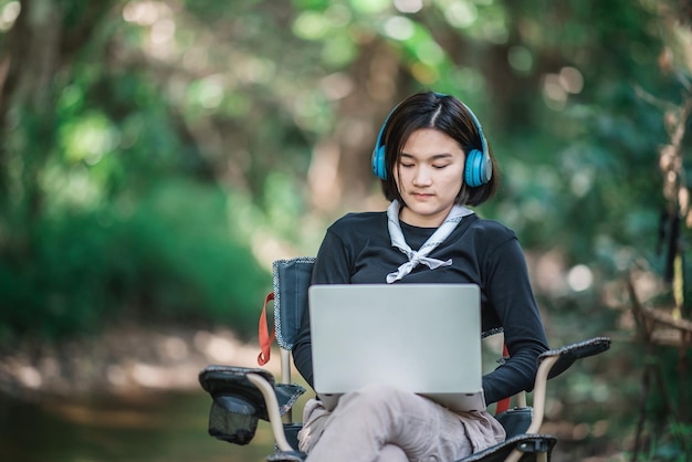 Feliz mujer joven sentada en una silla de camping usando auriculares escuchando música de una computadora portátil mientras se relaja acampando en el bosque