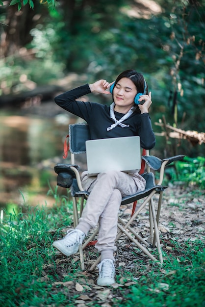 Feliz mujer joven sentada en una silla de camping usando auriculares escuchando música de una computadora portátil mientras se relaja acampando en el bosque