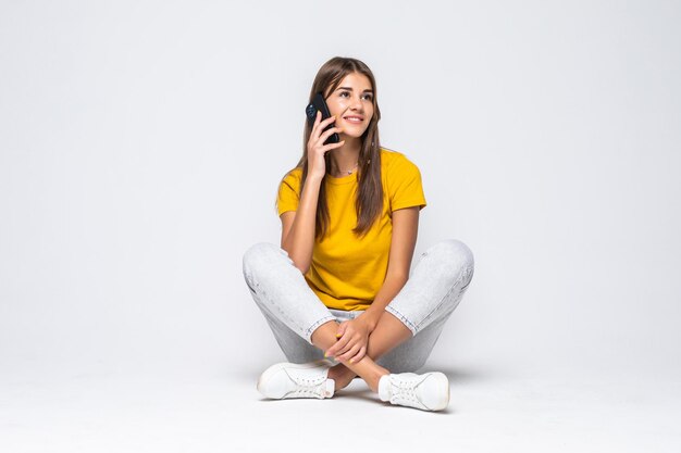 Feliz mujer joven sentada en el suelo hablando por un teléfono móvil aislado en blanco