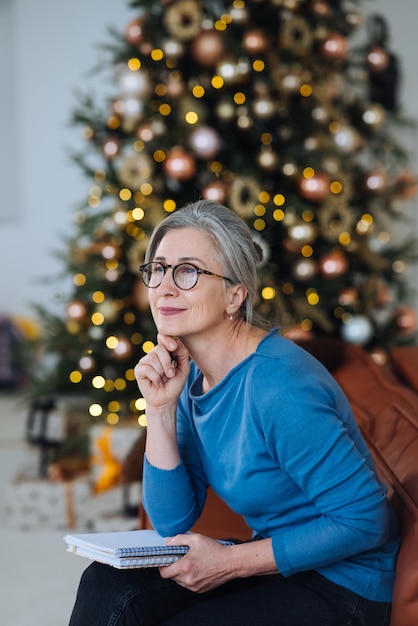 Feliz mujer mayor en gafas pensando y escribiendo en un diario en casa