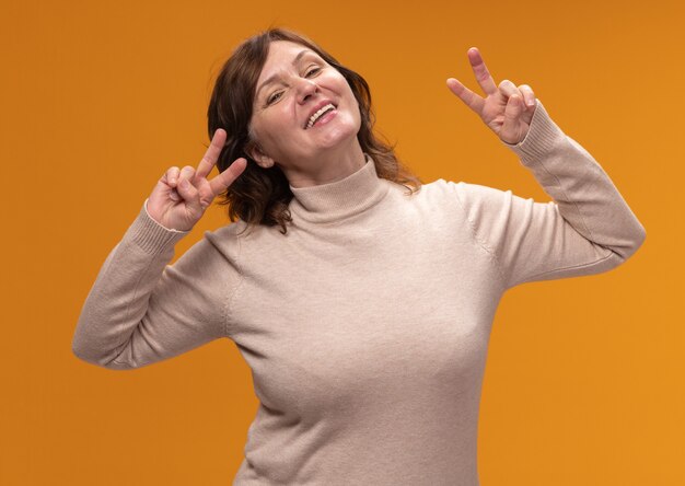 Feliz mujer de mediana edad en cuello alto beige sonriendo alegremente mostrando v-sign de pie sobre la pared naranja