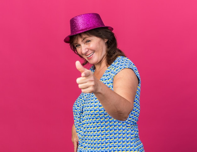 Feliz mujer de mediana edad con gorro de fiesta apuntando con el dedo índice sonriendo alegremente celebrando la fiesta de cumpleaños de pie sobre la pared rosa