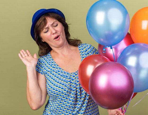 Feliz mujer de mediana edad con gorro de fiesta con un montón de globos de colores divirtiéndose celebrando la fiesta de cumpleaños de pie sobre la pared verde