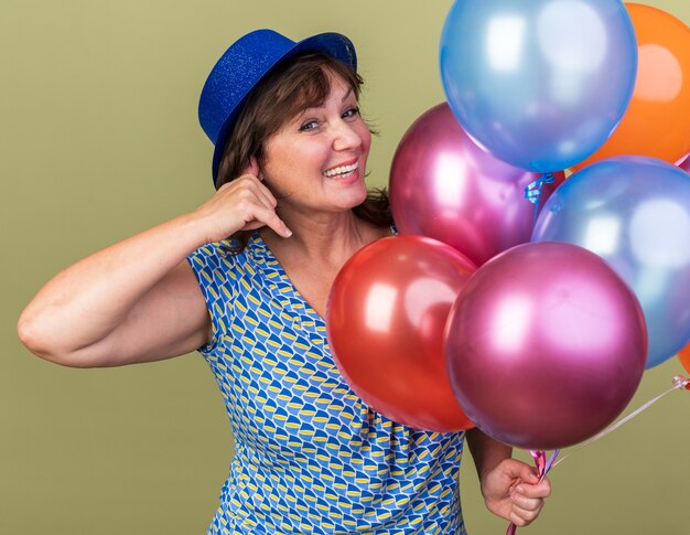 Feliz mujer de mediana edad con gorro de fiesta con un montón de globos de colores sonriendo alegremente haciendo gesto de llamarme