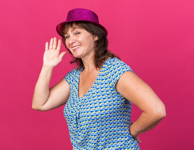 Feliz mujer de mediana edad con gorro de fiesta sonriendo alegremente saludando con la mano celebrando la fiesta de cumpleaños de pie sobre la pared rosa