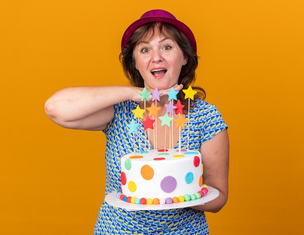 Feliz mujer de mediana edad con gorro de fiesta sosteniendo pastel de cumpleaños sonriendo alegremente