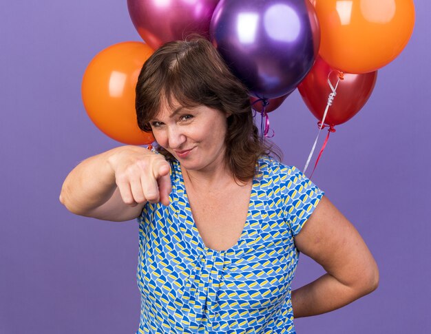 Feliz mujer de mediana edad con un montón de globos de colores apuntando con el dedo índice sonriendo alegremente celebrando la fiesta de cumpleaños de pie sobre la pared púrpura