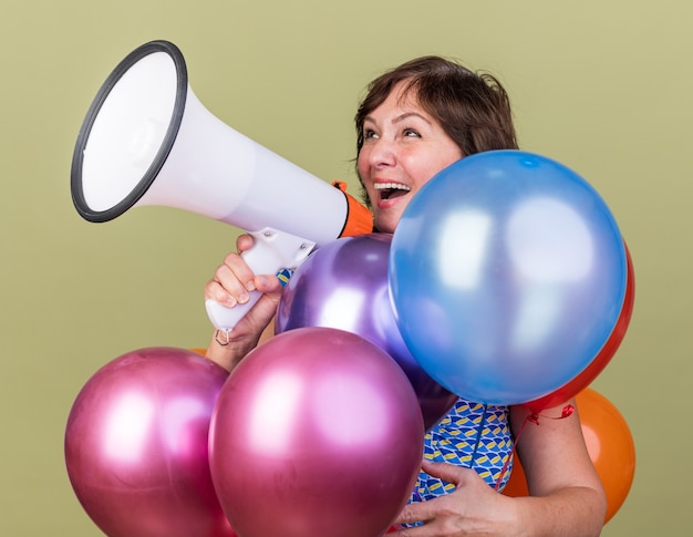 Feliz mujer de mediana edad con un montón de globos de colores gritando al megáfono