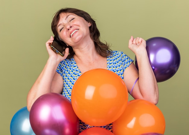 Feliz mujer de mediana edad montón de globos de colores sonriendo alegremente mientras habla por teléfono móvil