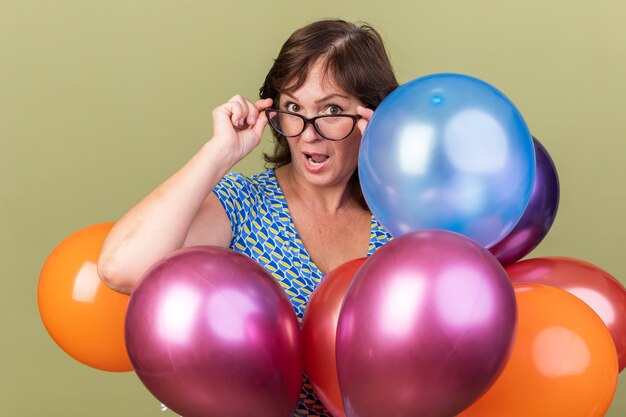 Feliz mujer de mediana edad en vasos con montón de globos de colores sorprendidos