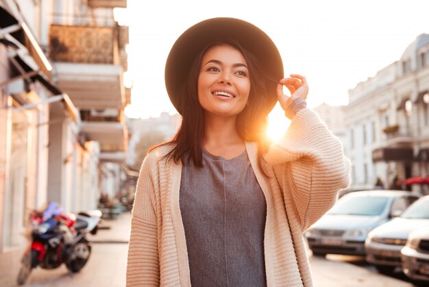 Feliz mujer moda asiática ajustando su sombrero mientras caminaba por la calle de la ciudad