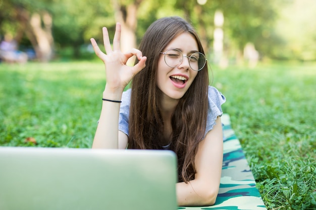 Feliz mujer morena en anteojos tumbado en la hierba en el parque con ordenador portátil y mostrando signo ok