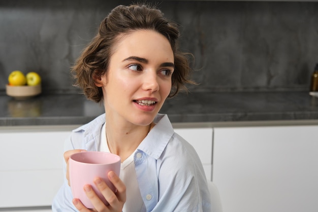 Feliz mujer morena bebiendo café en casa chica con taza rosa se sienta en su cocina y disfruta relajarse