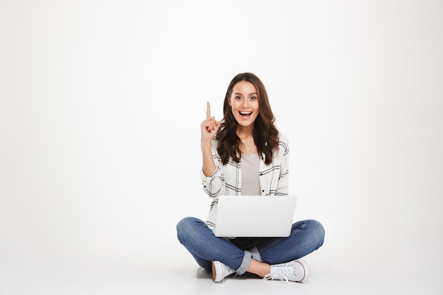 Feliz mujer morena en camisa sentada en el suelo con la computadora portátil mientras tiene idea y mirando a la cámara sobre gris