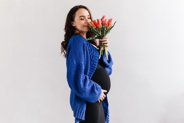 Feliz mujer morena en chaqueta de punto azul y vestido negro huele tulipanes. Encantadora dama embarazada tiene ramo de flores en aislados.