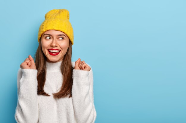 Feliz mujer morena levanta los puños cerrados, sonríe positivamente, mira a un lado, está de buen humor, celebra el examen aprobado con éxito en la universidad, usa poses de suéter blanco suelto en interiores