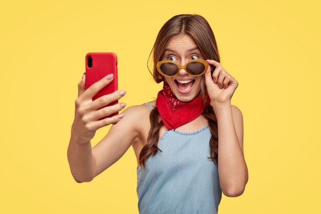 Feliz mujer morena con mirada alegre, se regocija comprando elegantes tonos para el verano, se prepara para las vacaciones, hace una foto de sí misma en el celular, modelos contra la pared amarilla. Chica toma selfie