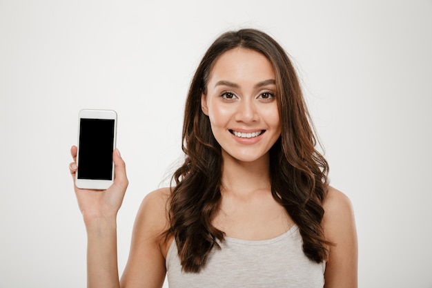 Feliz mujer morena mostrando la pantalla del teléfono inteligente en blanco y mirando a la cámara sobre gris