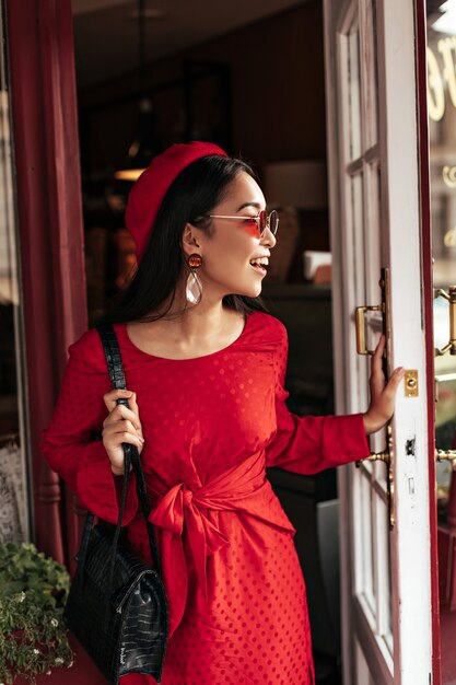 Feliz mujer morena optimista en elegante vestido rojo, boina de moda y gafas de sol sostiene bolso negro, sonríe y abre la puerta