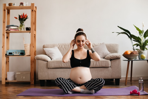 Foto gratuita feliz mujer morena en la parte superior escuchando música en auriculares en la alfombra de yoga púrpura