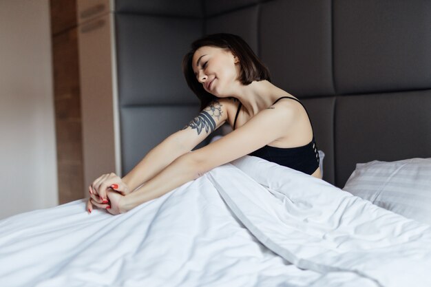 Feliz mujer morena de pelo largo en cama blanca en suave luz de la mañana bajo el edredón