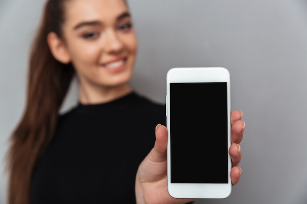Feliz mujer morena en ropa negra que muestra la pantalla del teléfono inteligente en blanco