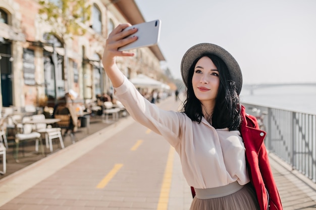 Feliz mujer morena en traje romántico haciendo selfie cerca de street cafe