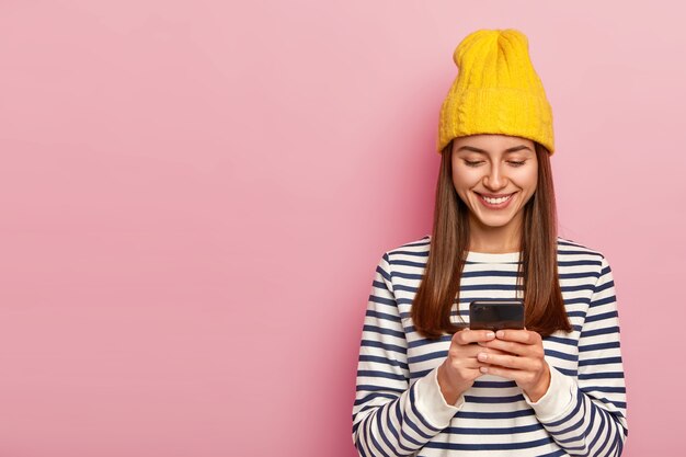 Feliz mujer morena usa sombrero amarillo, suéter de rayas casual, sostiene un teléfono celular moderno, verifica el buzón de correo electrónico, recibe un mensaje de un amigo en el extranjero