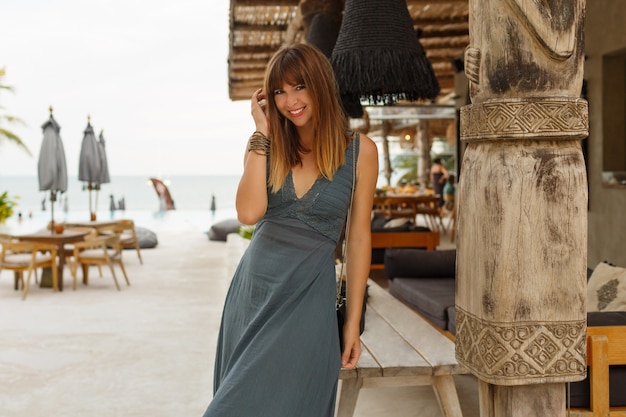 Feliz mujer morena en vestido sexy posando en un elegante restaurante de playa en estilo asiático.