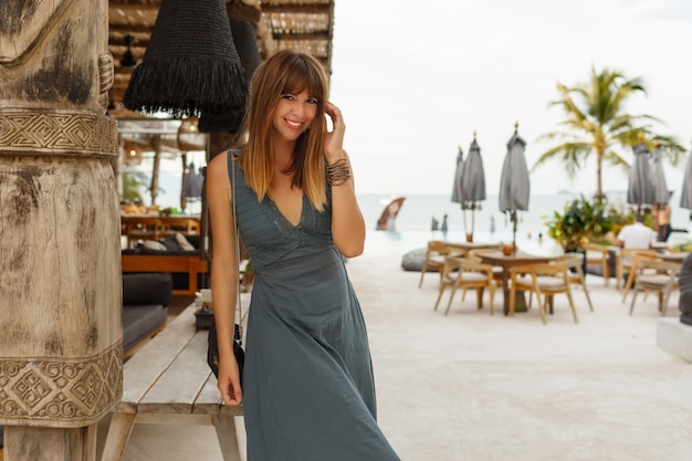 Feliz mujer morena en vestido sexy posando en un elegante restaurante de playa en estilo asiático.