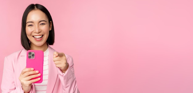 Feliz mujer de negocios asiática riéndose apuntándote con el dedo y grabando videos tomando fotos en un teléfono inteligente usando el fondo rosa del teléfono móvil