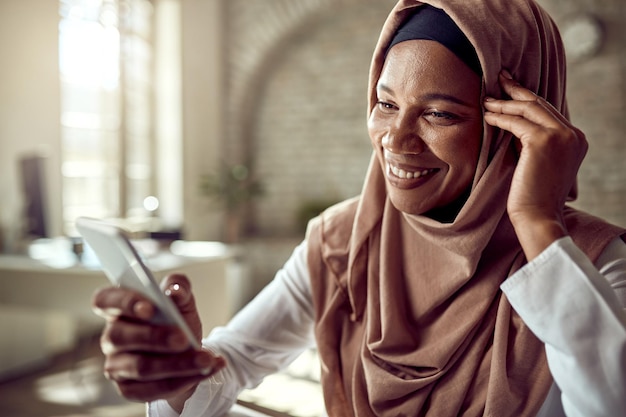 Feliz mujer de negocios islámica negra usando teléfono celular y mensajes de texto mientras trabaja en la oficina