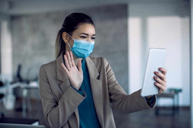 Feliz mujer de negocios con mascarilla ondeando durante una videollamada a través del panel táctil en la oficina