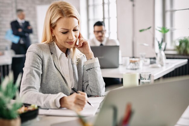 Feliz mujer de negocios trabajando en su escritorio y tomando notas en su bloc de notas Hay gente en el fondo