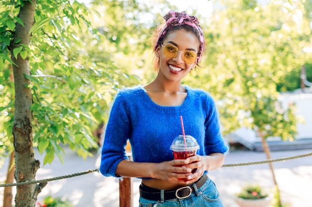 Feliz mujer negra con elegante peinado con diadema pasar su fin de semana en el parque, caminar y beber limonada.