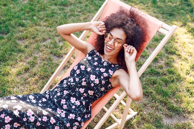 Feliz mujer negra de moda con elegante peinado rizado sentado en chaise-lounge en increíble césped verde