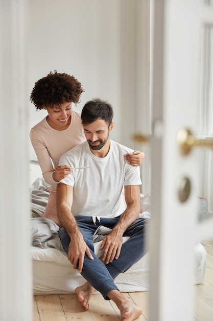 Foto gratuita feliz mujer negra muestra la prueba de embarazo al esposo, satisfecha con el resultado positivo, posa en el dormitorio del piso moderno, se regocija con las buenas noticias, lista para convertirse en padres. pareja familiar interior. paternidad